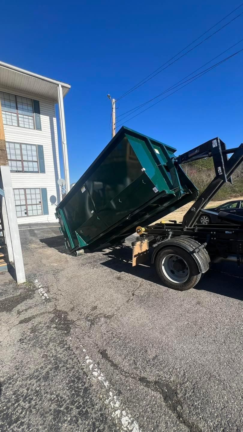 A green dumpster tipped on a truck lift beside a white building on a gravel lot.