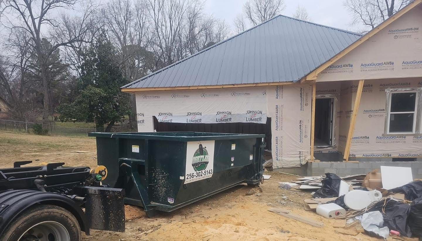 Green dumpster and truck at a construction site beside a partially built house with debris nearby