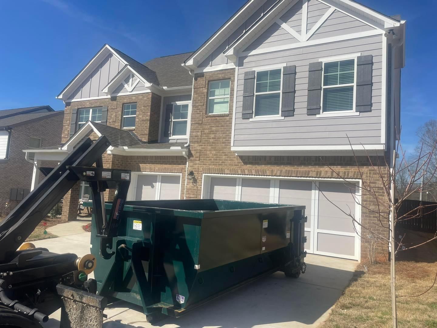 Large dumpster and black truck bed in a suburban driveway in front of a two-story house with stone siding.