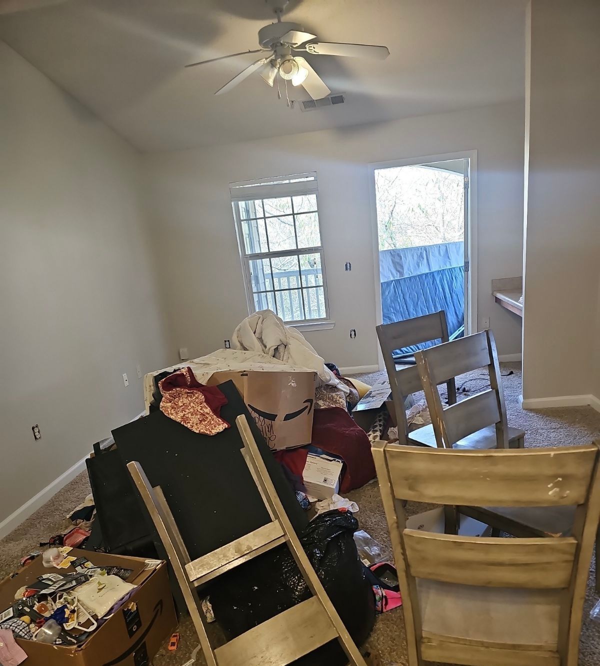 Cluttered room with overturned furniture, boxes, and a ceiling fan near a glass door.
