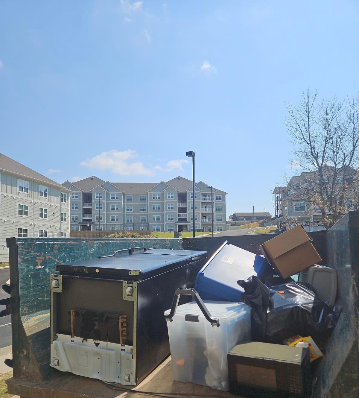 Overflowing trash bins and scattered cardboard in a fenced apartment courtyard under a bright blue sky