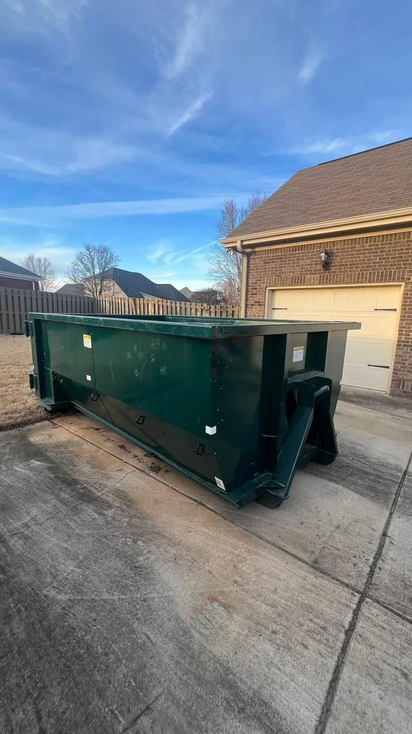 Green dumpster on a driveway beside a brick garage under a blue sky.