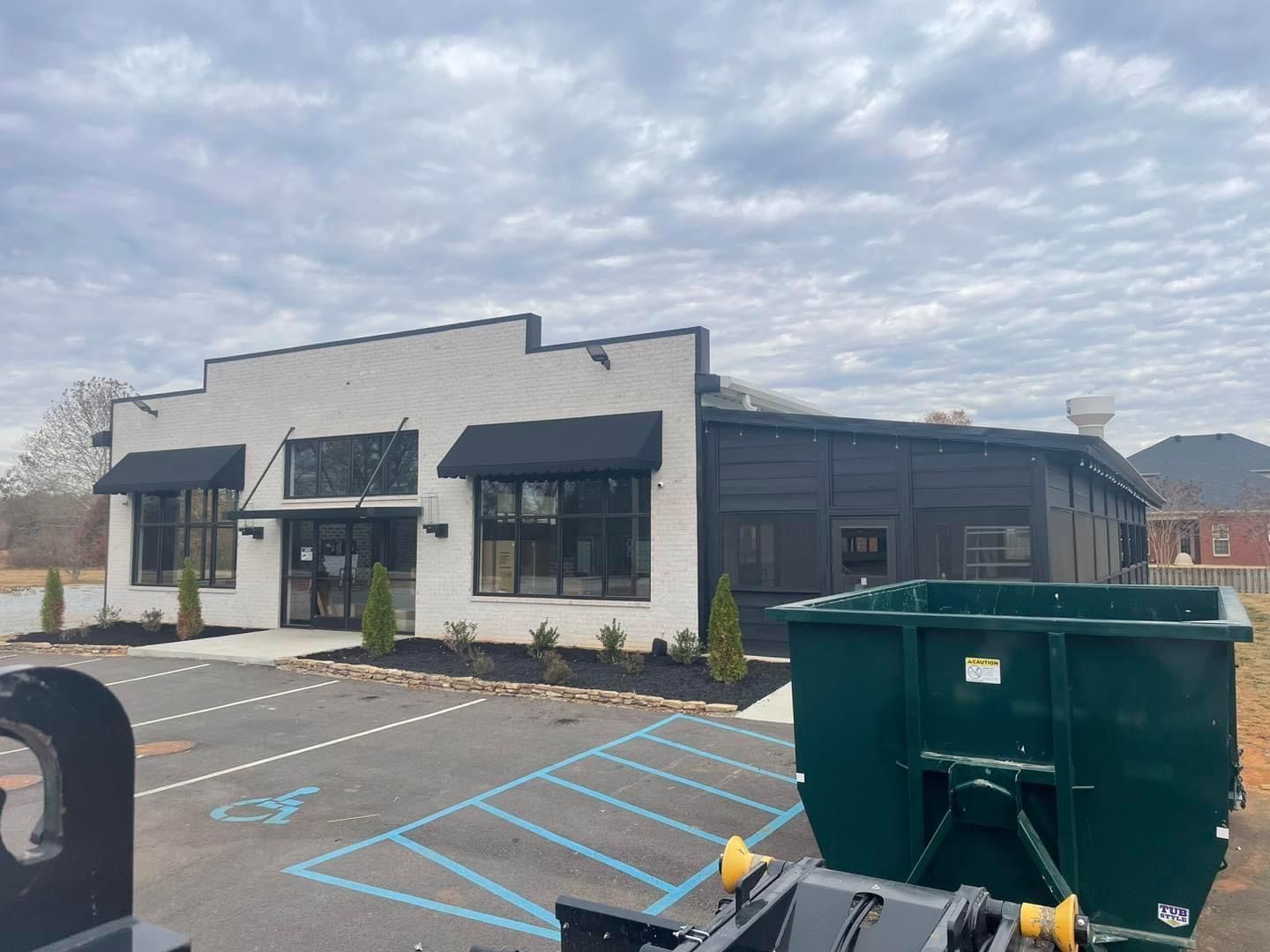 Commercial building with black awnings, empty accessible parking spaces, and green dumpsters in front.