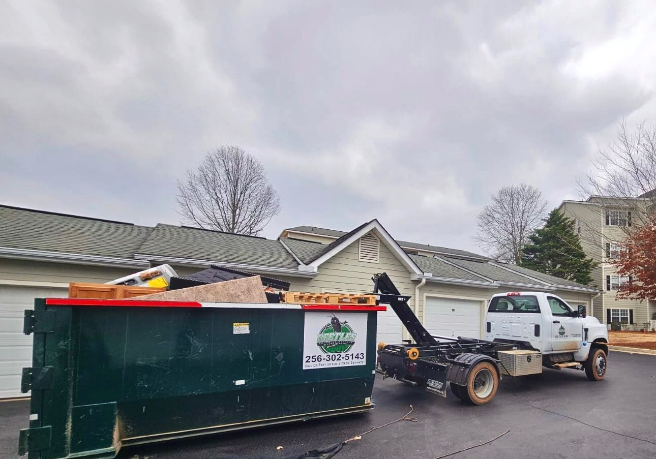 Tow truck hauling a white pickup beside a green dumpster in a residential street under cloudy skies