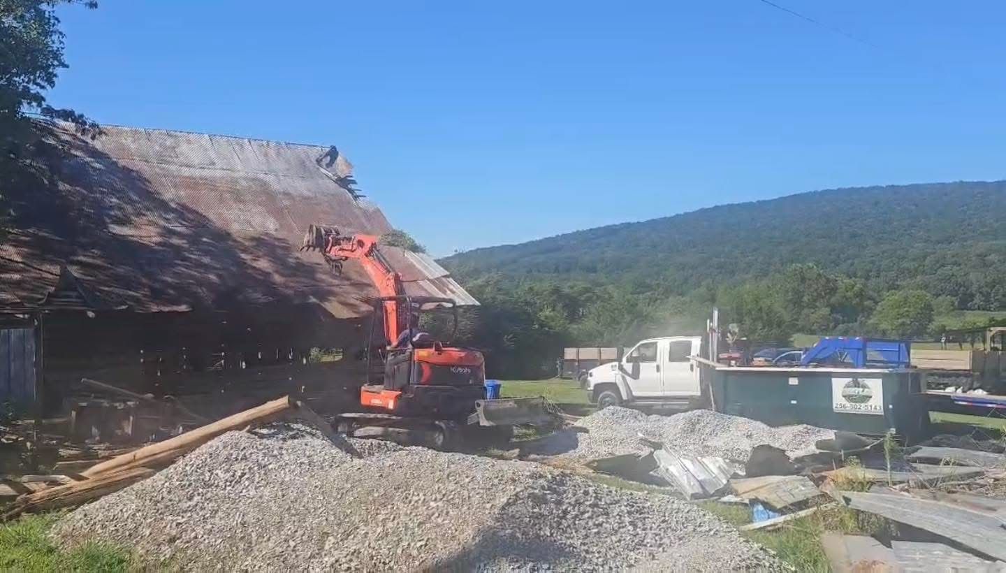 Construction machinery clearing rocks beside a dirt road in a mountain valley, with a white truck nearby.