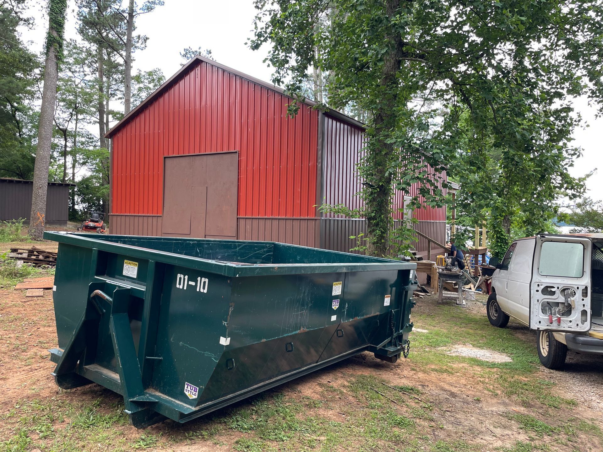 Large green dumpster in front of a red barn in a wooded yard, with a white truck at right.