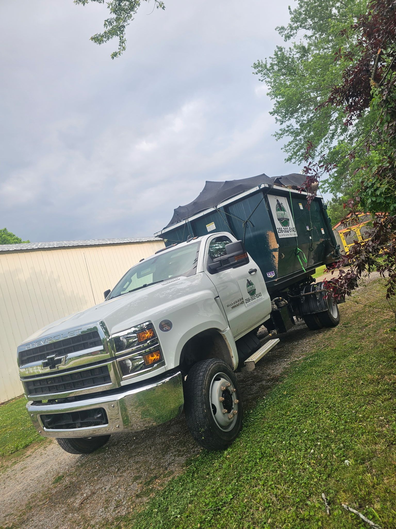Silver pickup truck on a gravel drive beside a black dump trailer, with trees and a cloudy sky.