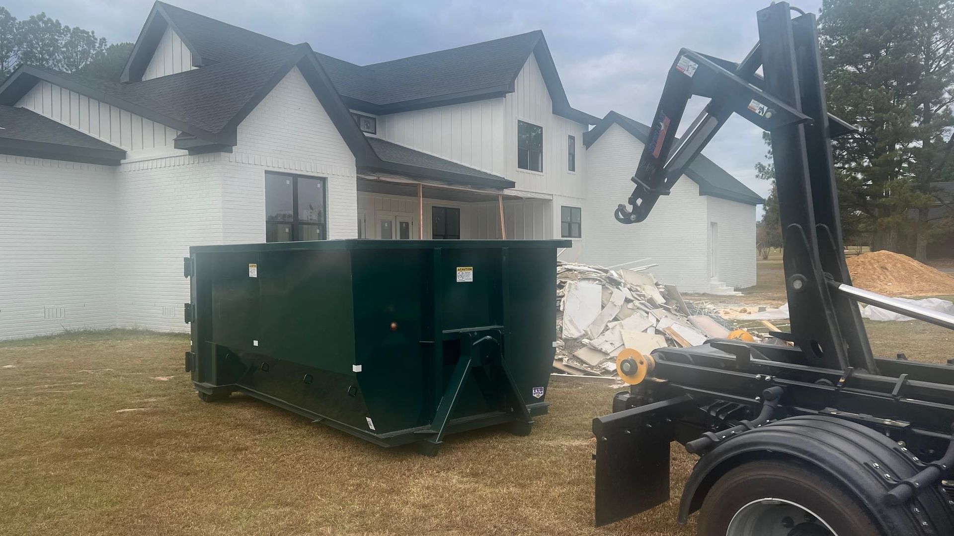 Green dumpster and excavator beside a house under construction on a dirt lot