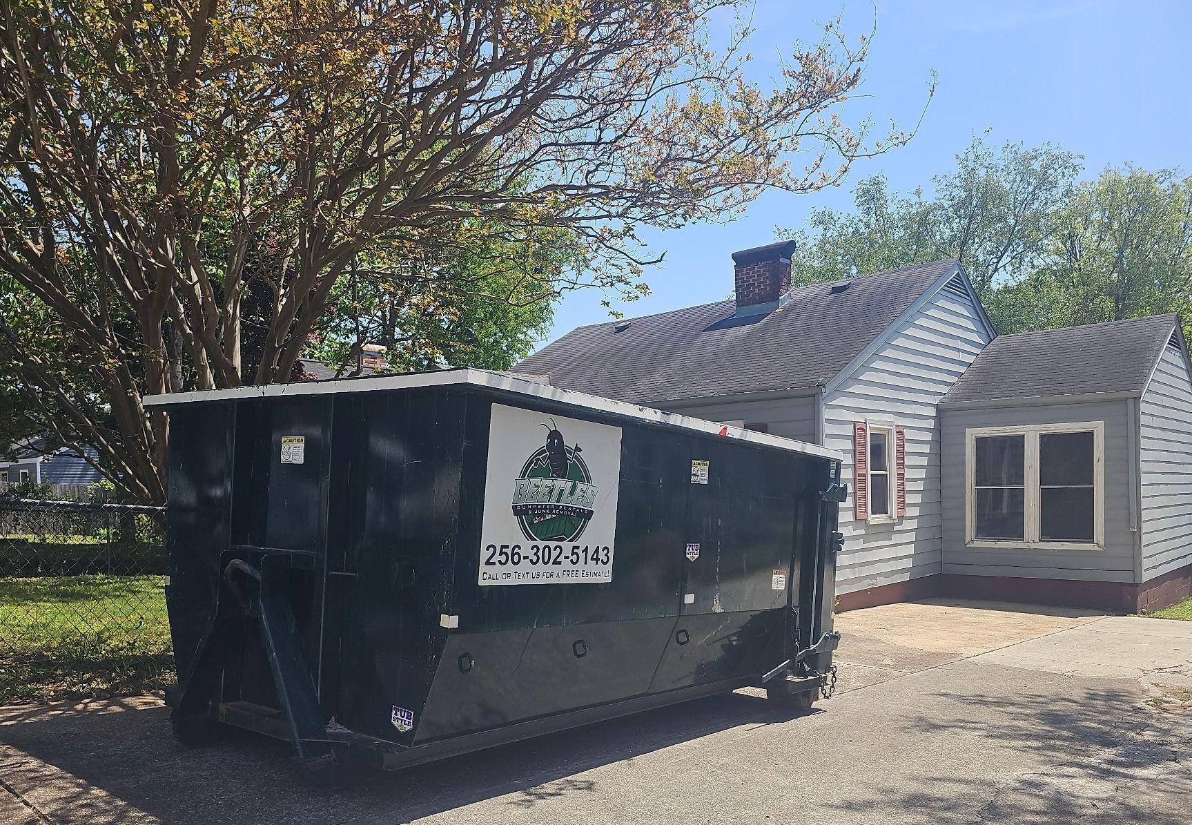 Black dumpster in front of a small gray house with white trim and trees in sunlight