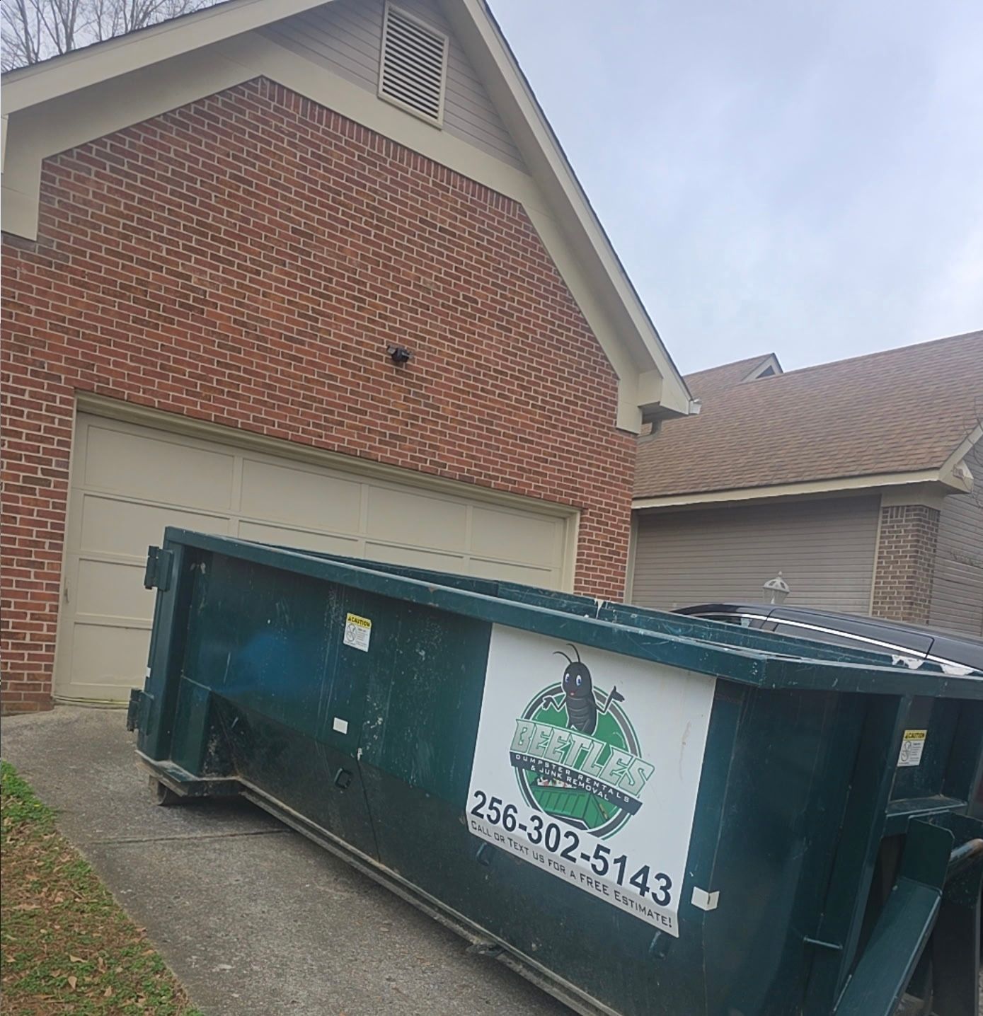 Large green dumpster in front of a brick house garage, with a white logo and phone number on the side