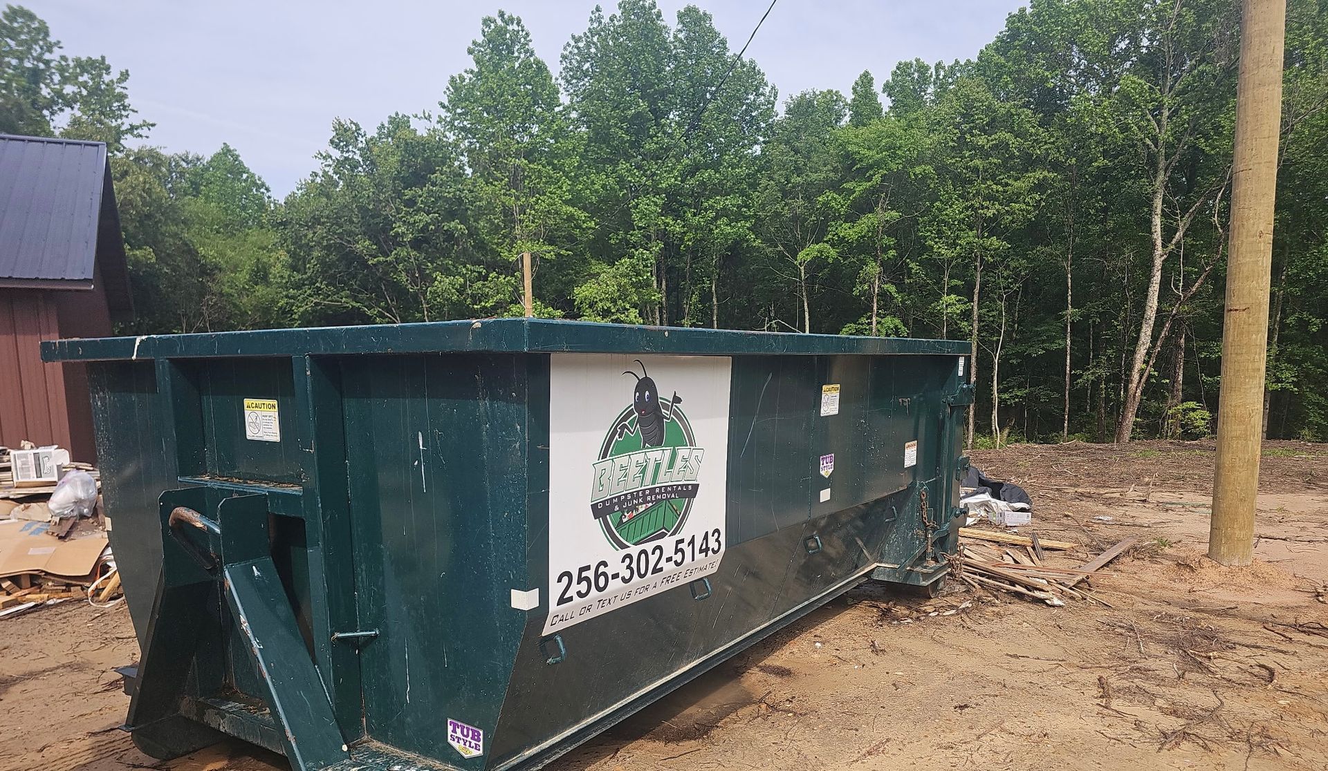 Green dumpster at a construction site beside trees and a building.