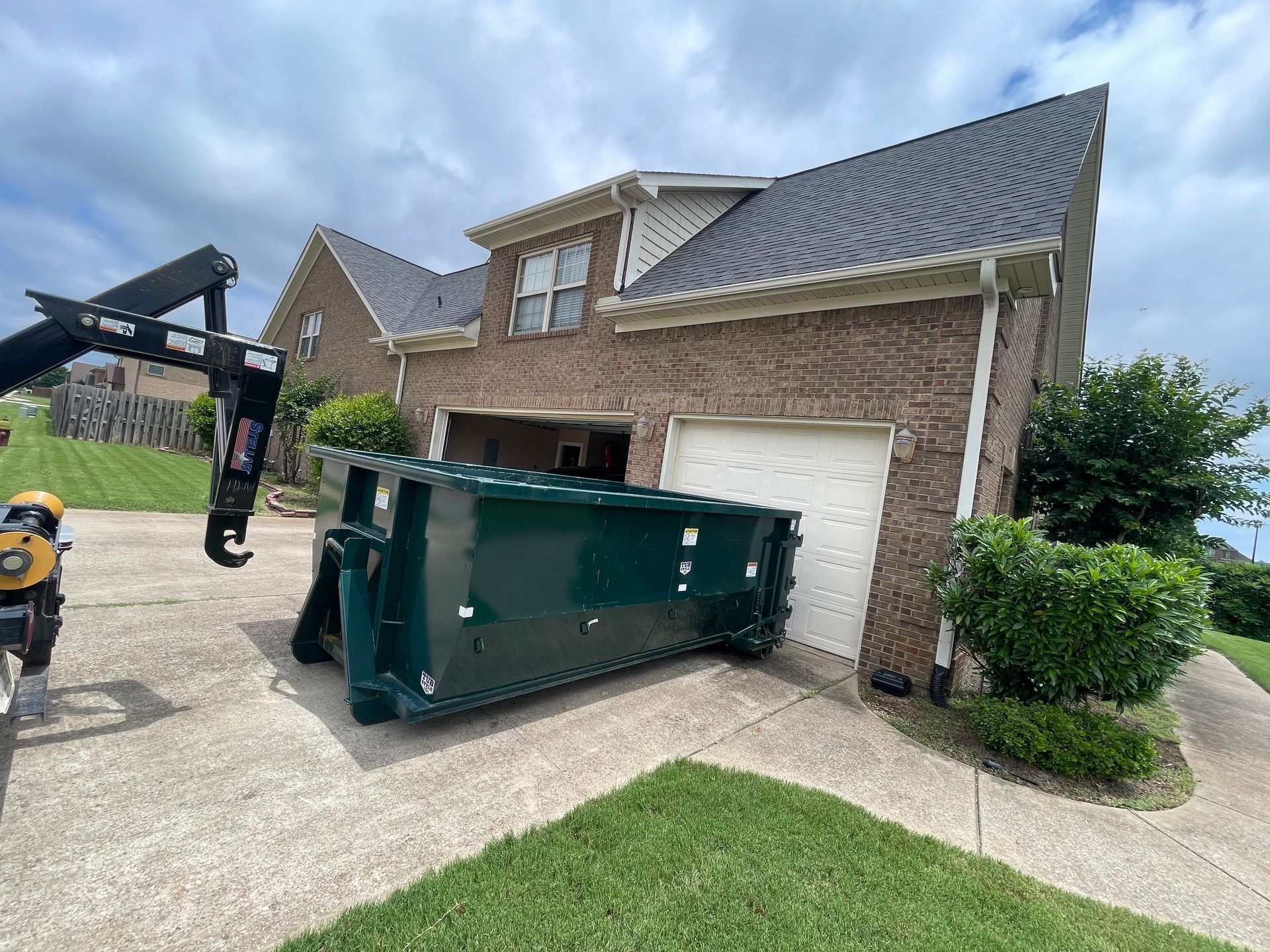 Dumpster being loaded onto a truck beside a brick house garage driveway