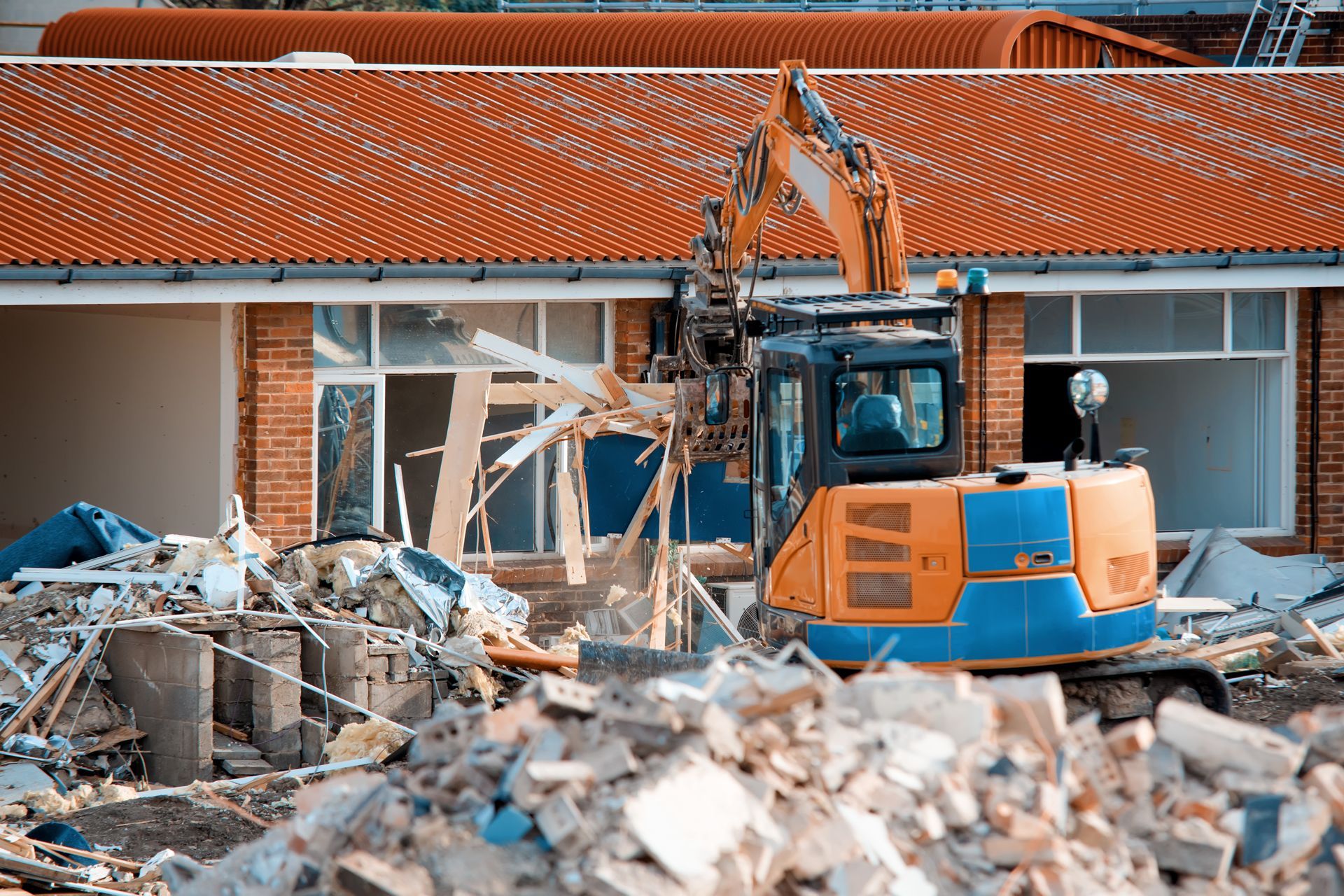 Excavator demolishing a house with a red-tiled roof, with rubble piled in front.