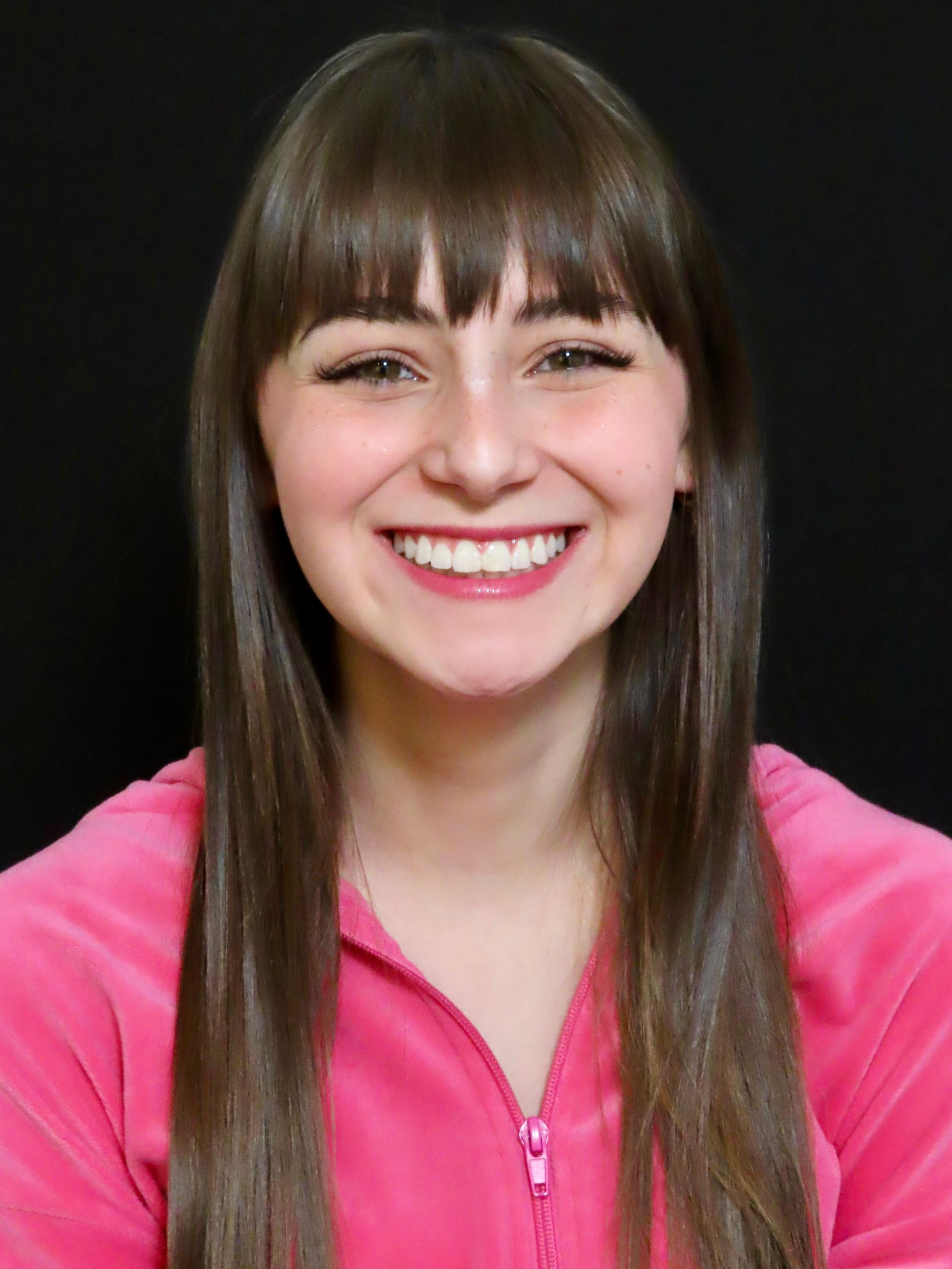 Smiling person with long brown hair wearing a pink zip-up top against a black background