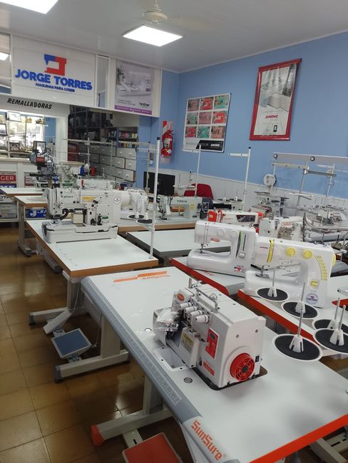 Taller de costura con filas de máquinas industriales sobre mesas y suministros de tela bajo luces brillantes.