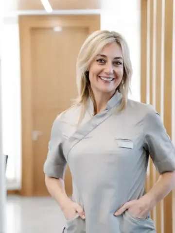 A woman is standing in a hallway with her hands in her pockets and smiling.
