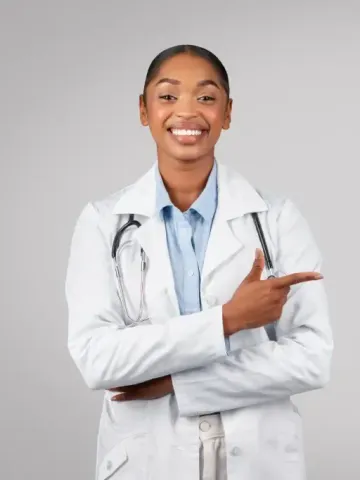 A woman in a lab coat with a stethoscope around her neck is smiling and pointing.