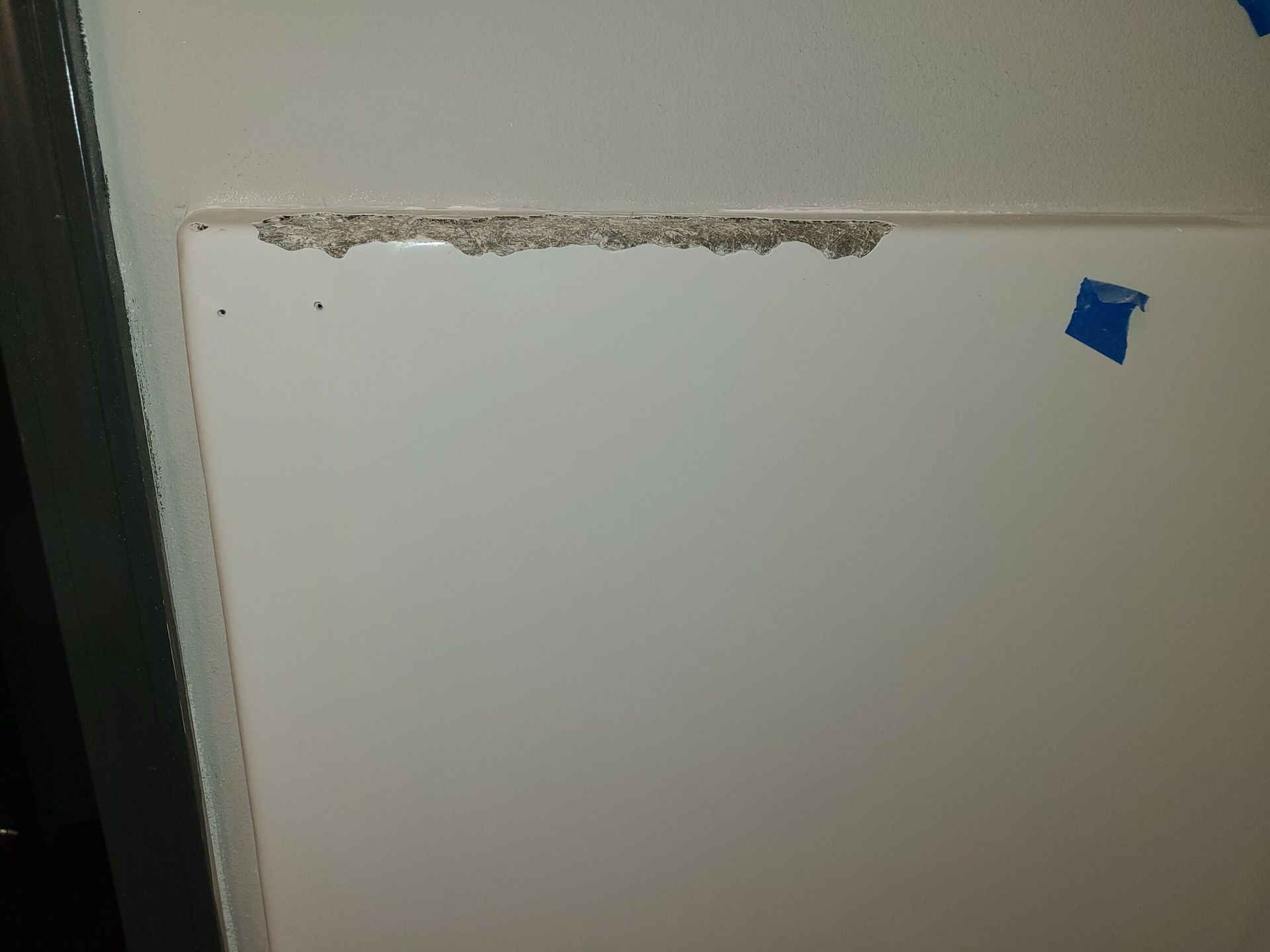 White wall or door panel with chipped paint along the top edge and a small blue tape square in the upper right.