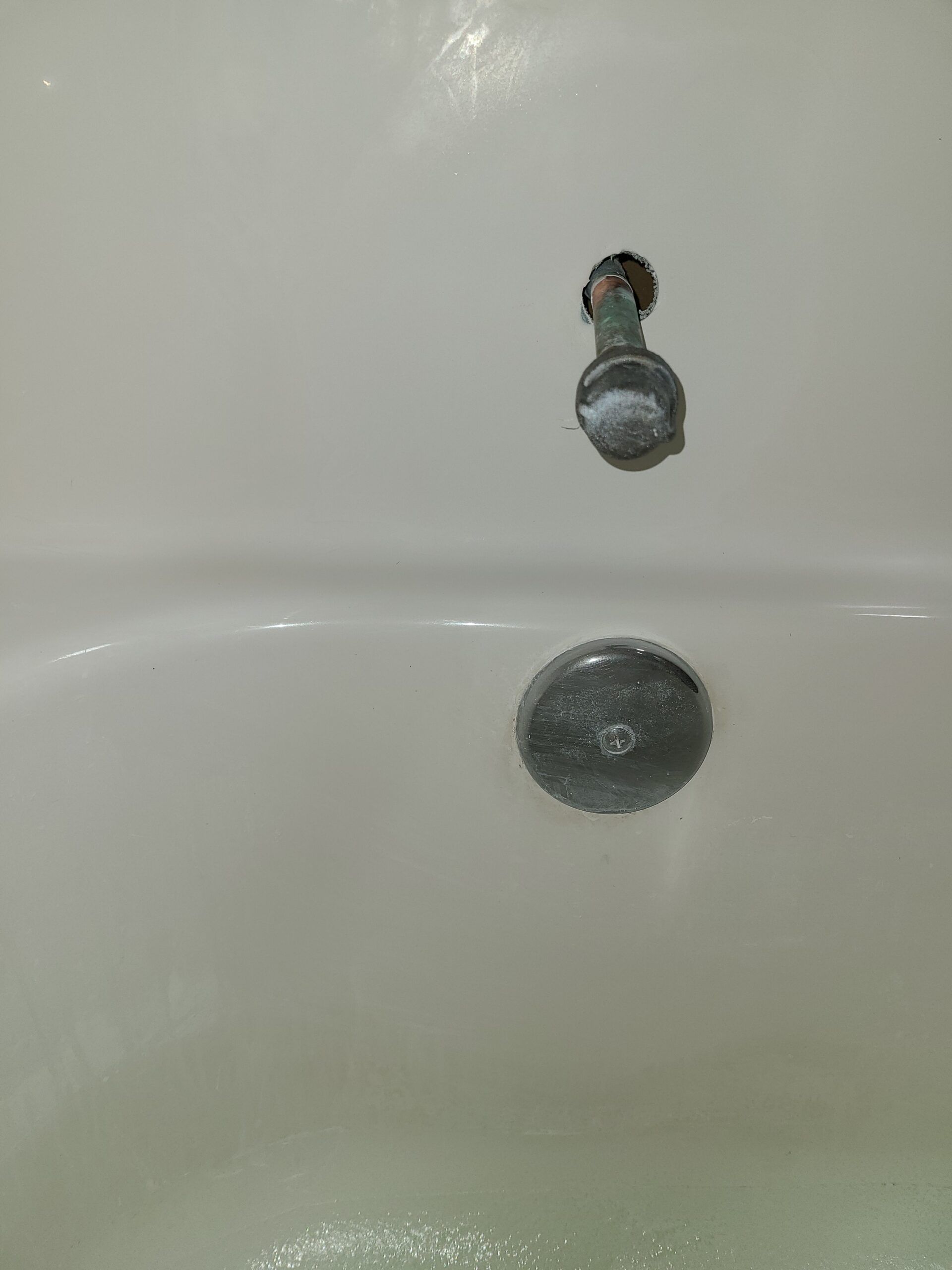 Close-up of a white bathtub drain and overflow opening with a small chrome faucet fixture above it