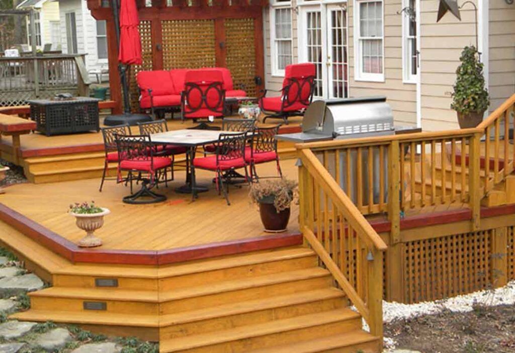Wooden backyard deck with red patio furniture, a grill, and stairs leading to a stone patio.