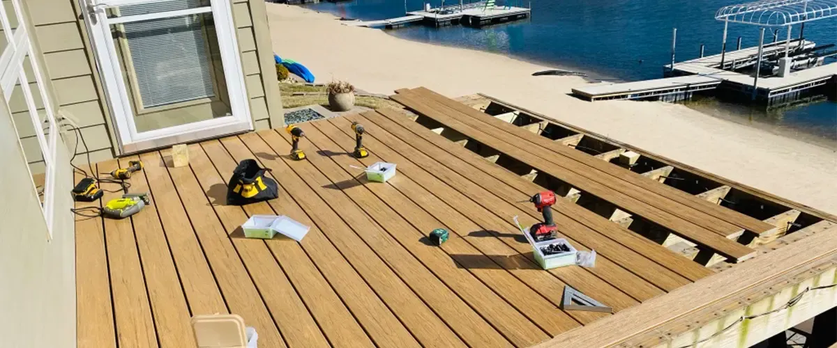 Tools and construction materials are scattered on a partially installed wooden deck overlooking a sandy beach and water.