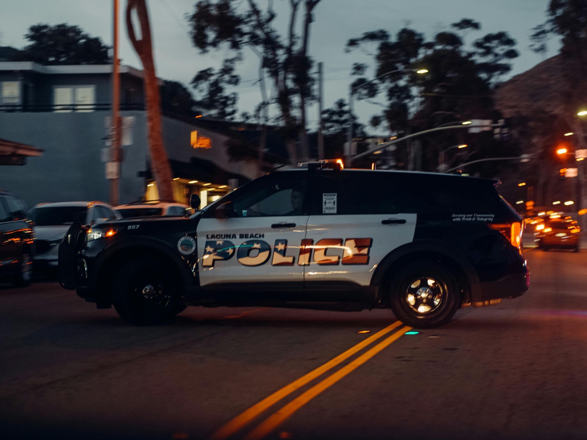 A black and white Laguna Beach police SUV driving across a street intersection during twilight.