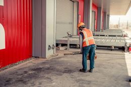 Worker in orange safety vest and helmet inspecting equipment beside a red industrial building.