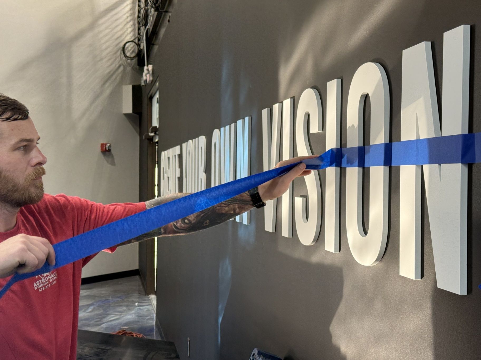 Man in red shirt uses blue strap to center letter on wall. White letters spell 