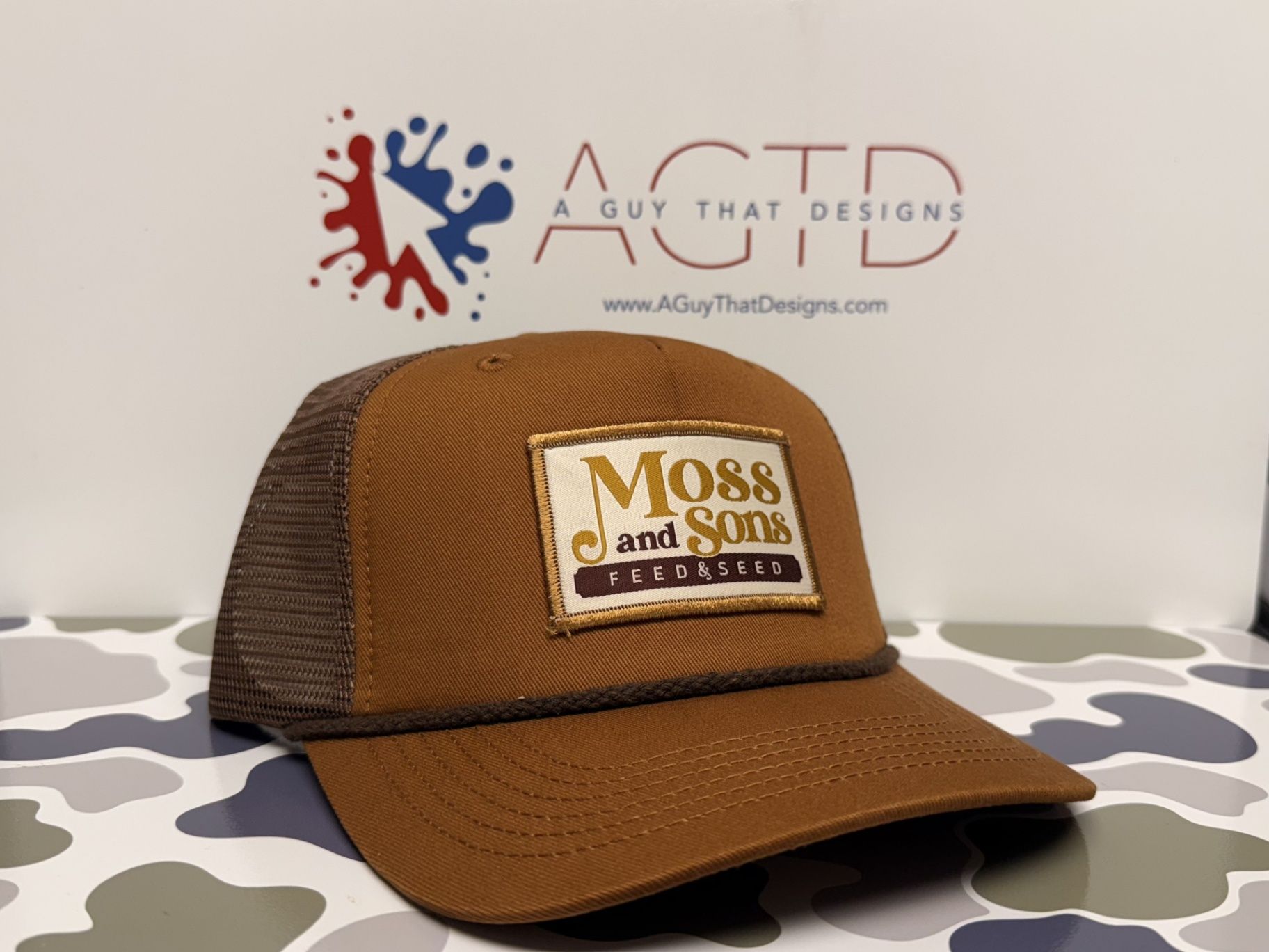 Brown and tan Moss and Sons Feed & Seed baseball cap with brown mesh backing, on camouflage.