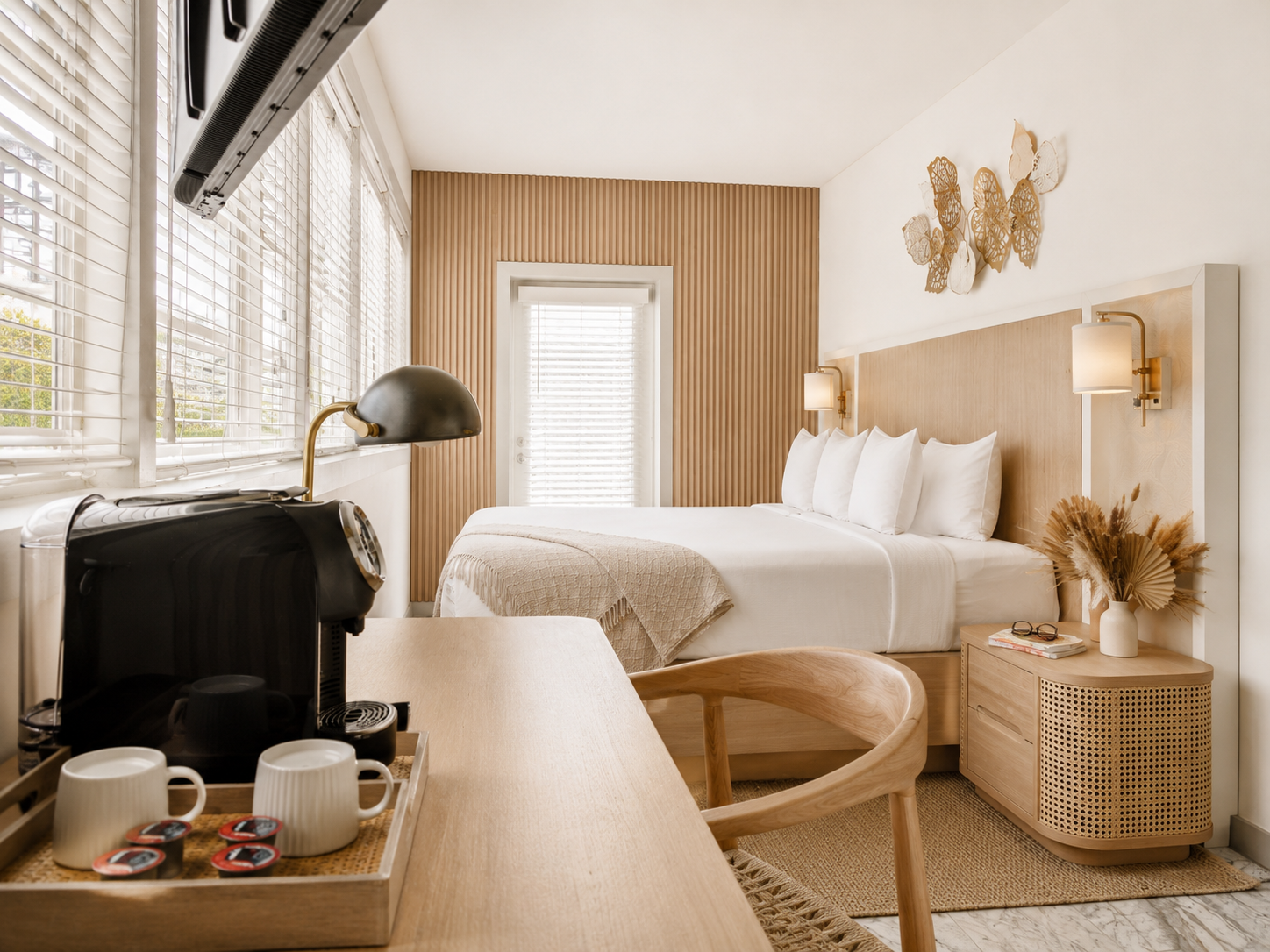 Bright bedroom with a bed, woven chair, wooden desk, and large windows overlooking trees