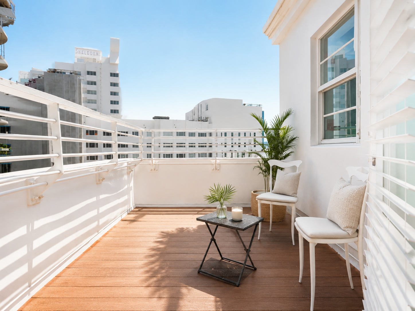 Sunny rooftop balcony with white chairs, a small table, potted plants, and city buildings beyond the railing