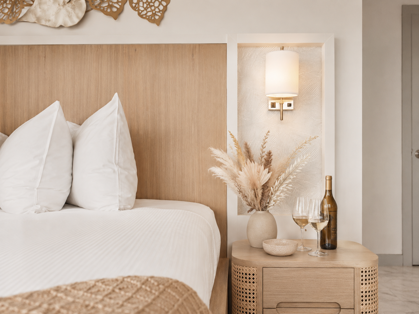 Neutral bedroom with white bedding, wood headboard, woven nightstand, lamp, and dried flowers