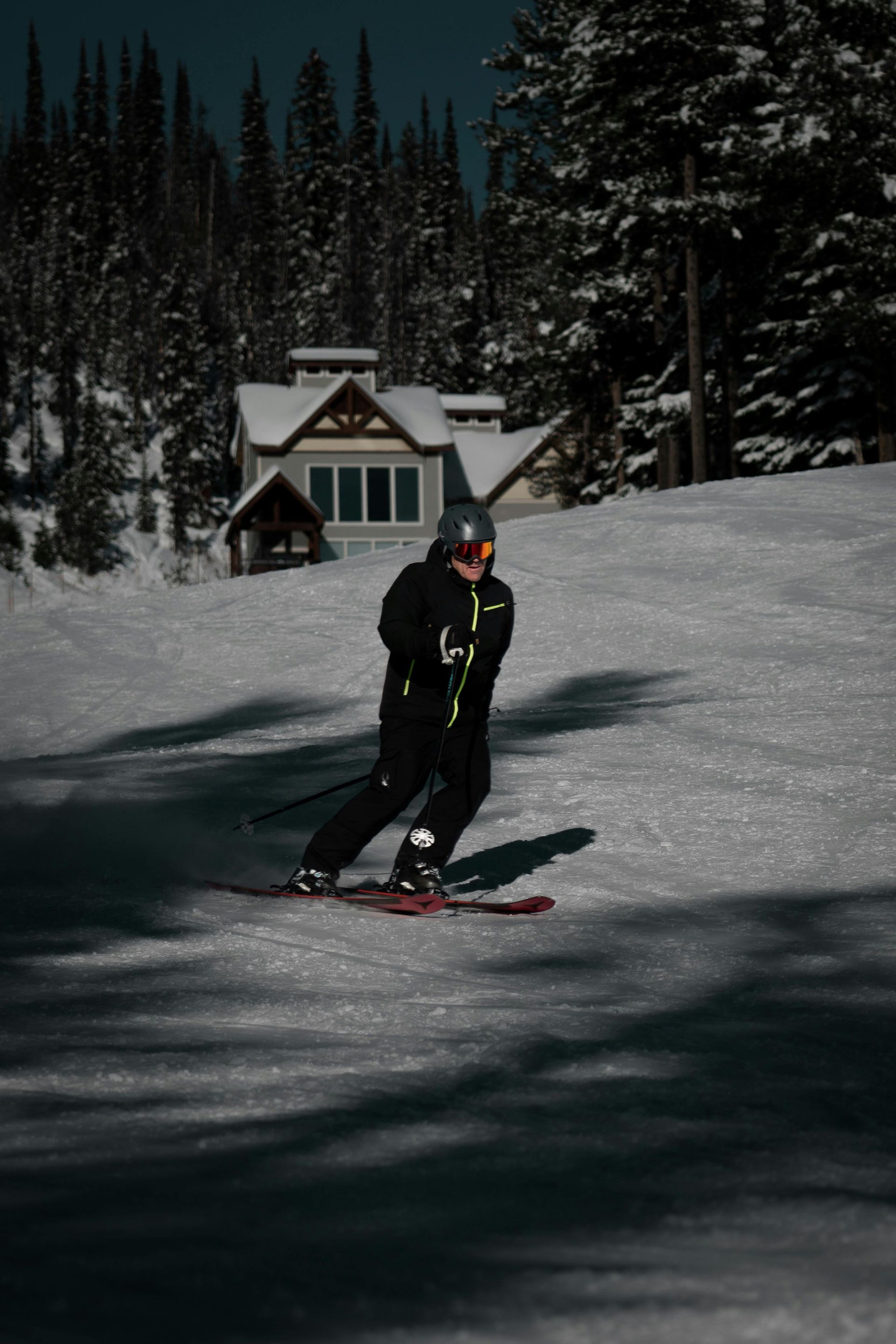 Skier in black on a snowy slope near a cabin and pine trees at dusk