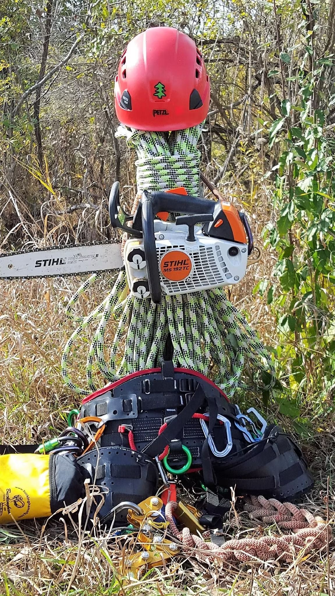 A bright red helmet rests on a coiled rope figure holding a white and orange chainsaw, with gear scattered on the ground.