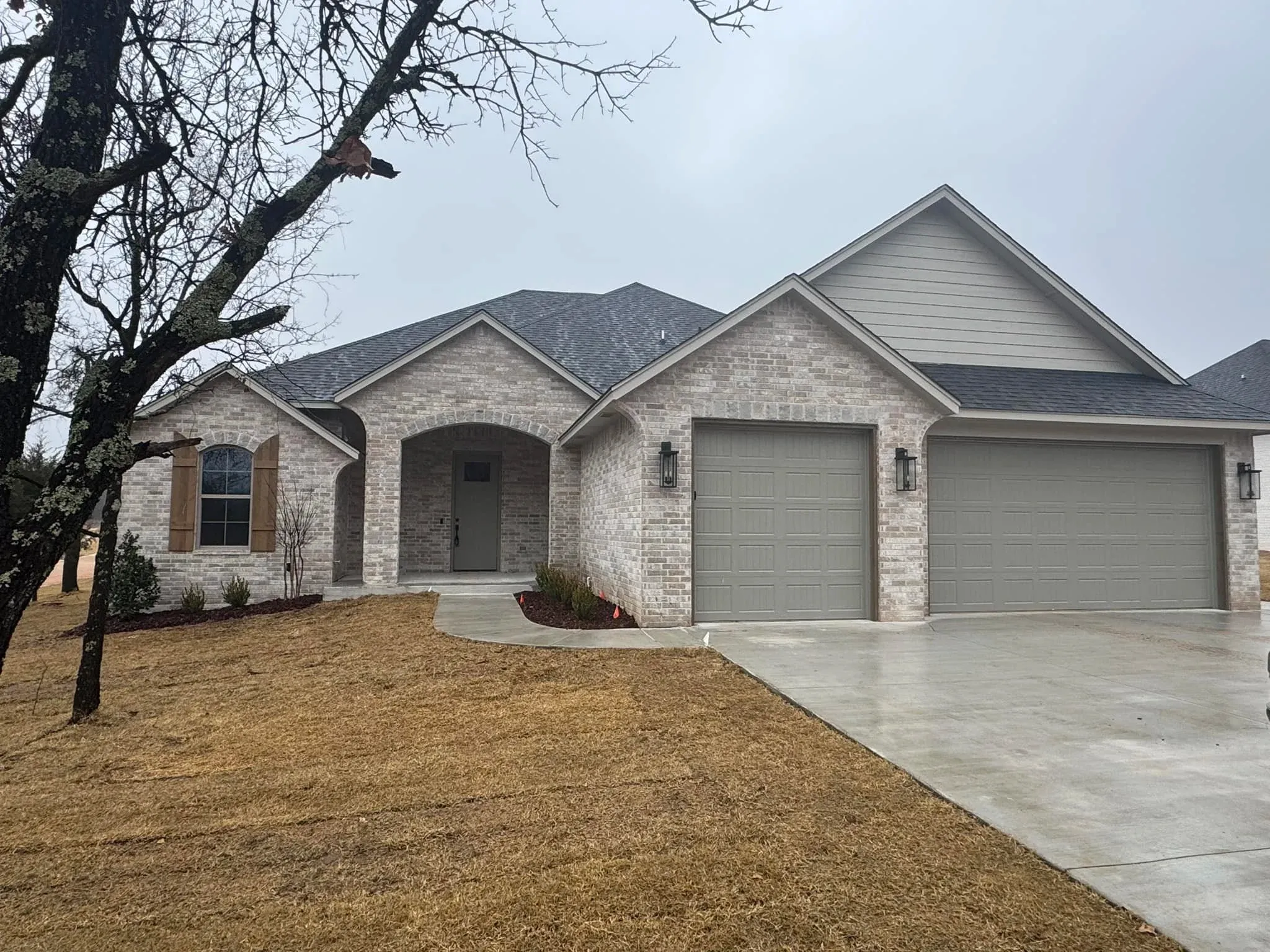 Gray brick house with two-car garage, under an overcast sky, with brown grass.