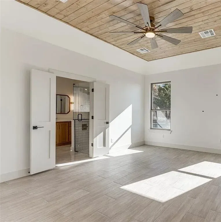 Bright bedroom with wood ceiling and white walls, leading to a bathroom. Sunlight streams in.