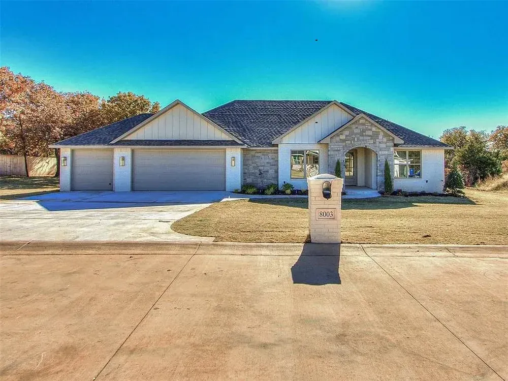 Modern single-story house with gray roof, stone accents, three-car garage, and mailbox on a sunny day.