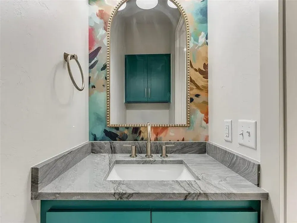 Teal bathroom vanity with marble countertop, floral wallpaper, and arched gold mirror.