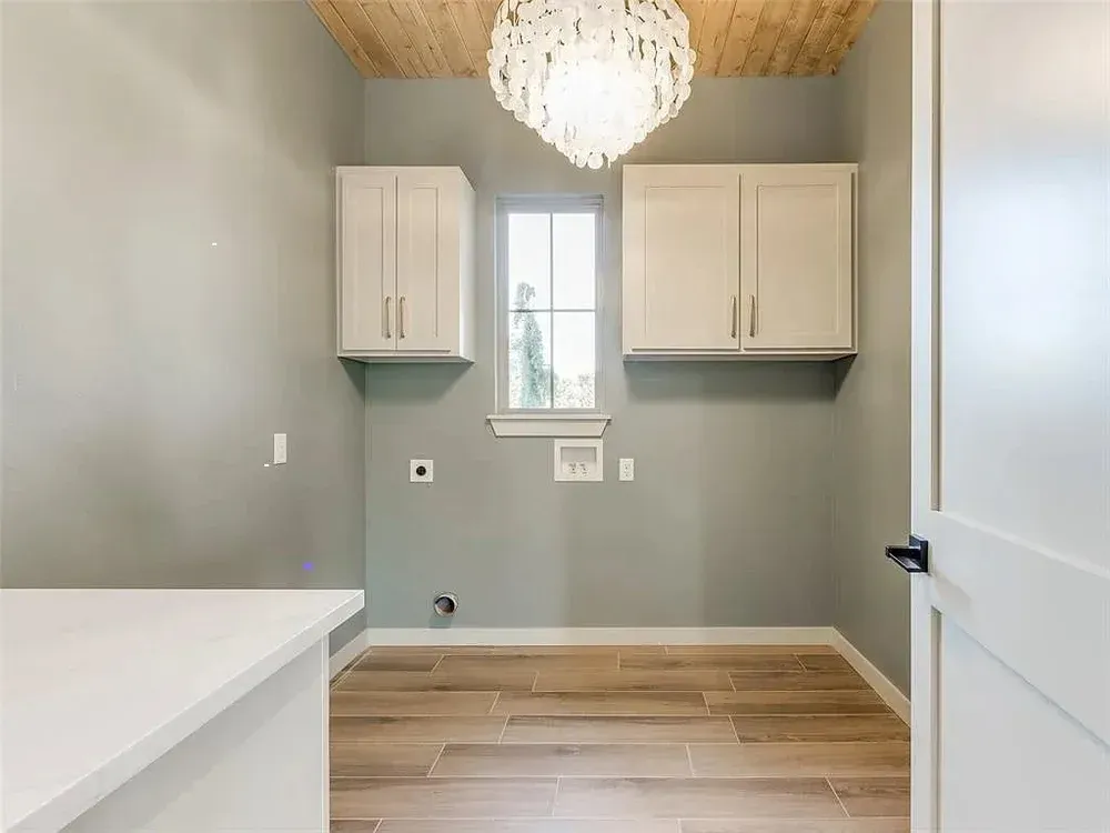 Laundry room with gray walls, wood floor, cabinets, window, and chandelier.