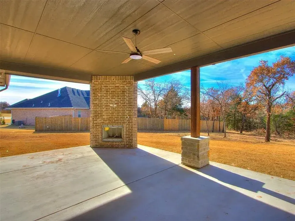 Covered patio with brick fireplace, ceiling fan, concrete floor, overlooking backyard and blue sky.