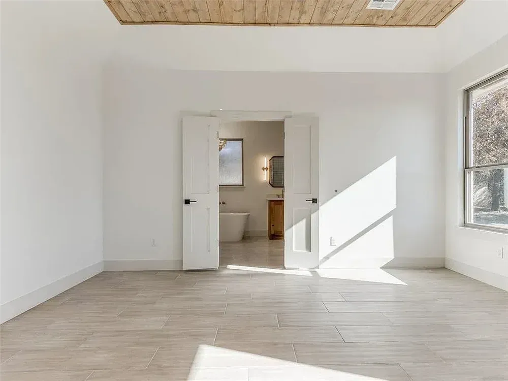 Empty room with open doors to a bathroom; natural light, neutral colors, wooden ceiling.