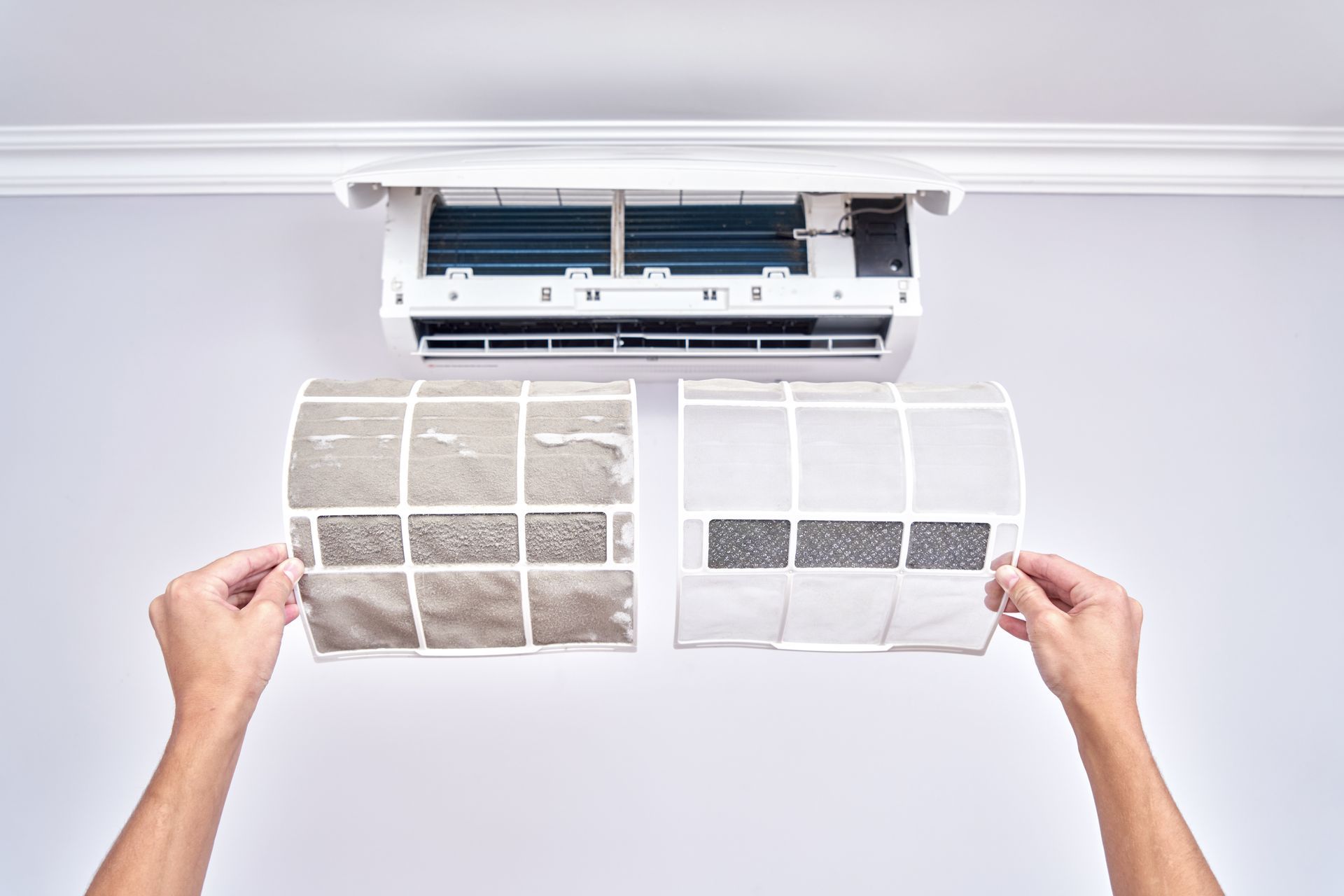 Hands holding a dirty air conditioner filter next to a clean one in front of a wall-mounted AC unit.