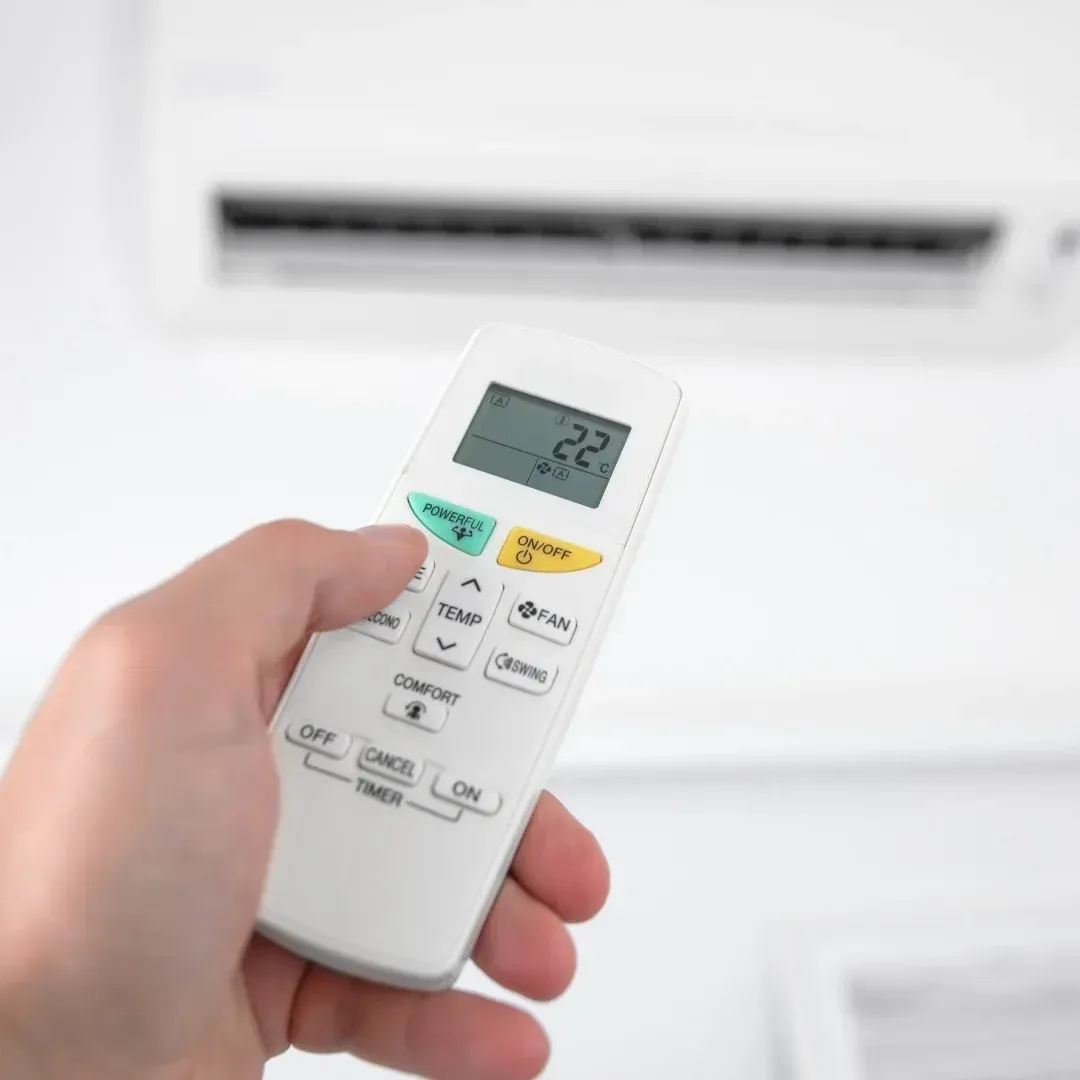 Hand holding a white air conditioner remote pointed at a wall-mounted AC unit