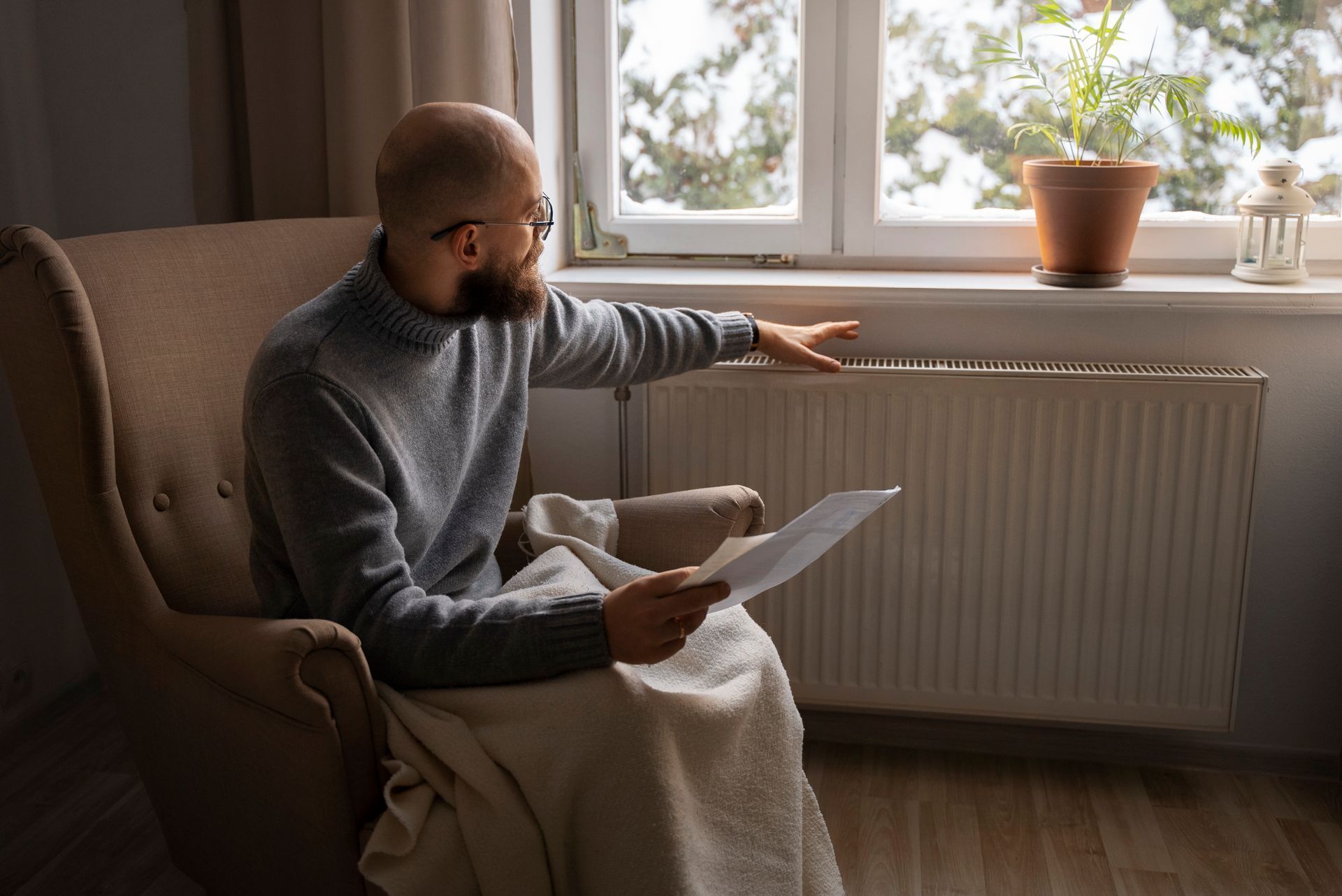 A person in a sweater and blanket sitting in a chair, holding a document and checking the temperature of a radiator.