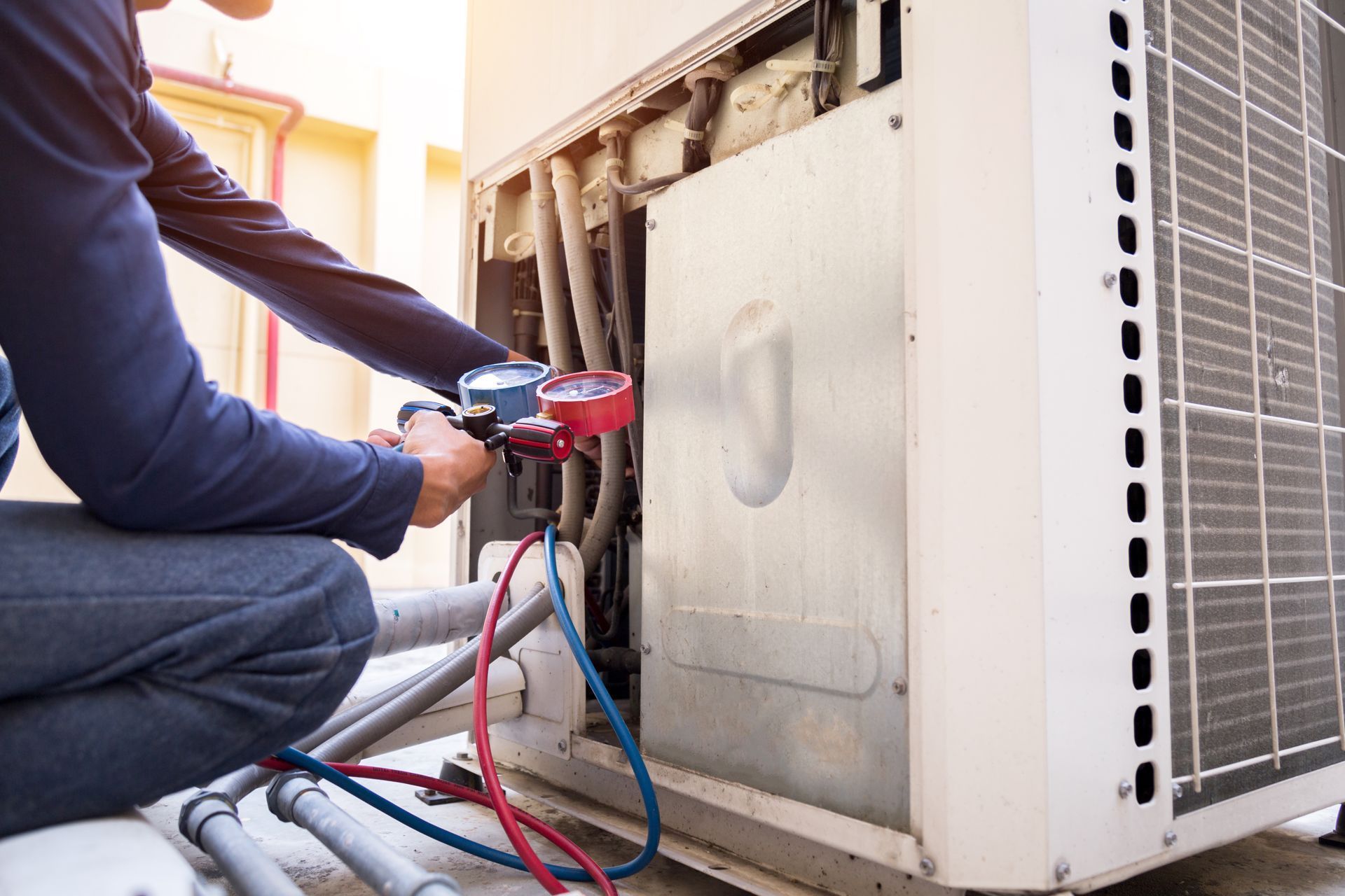 A technician in a blue shirt uses a manifold gauge set to service an outdoor air conditioning unit.
