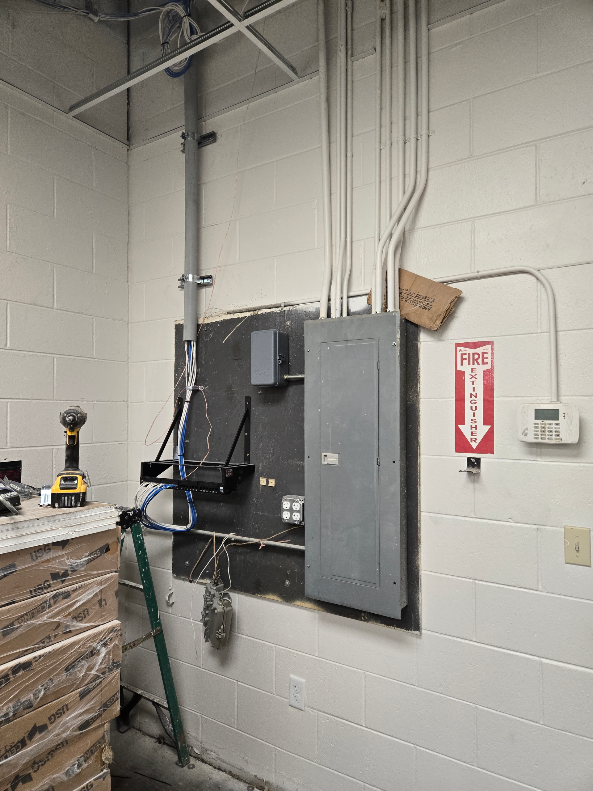 Utility room wall with electrical panels, conduit, and a red fire extinguisher sign above a workbench.