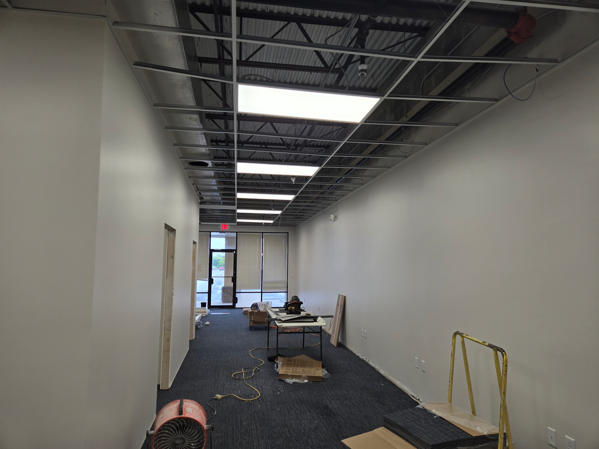 Long, unfinished hallway with exposed ceiling, white walls, carpet tile, and construction materials scattered inside