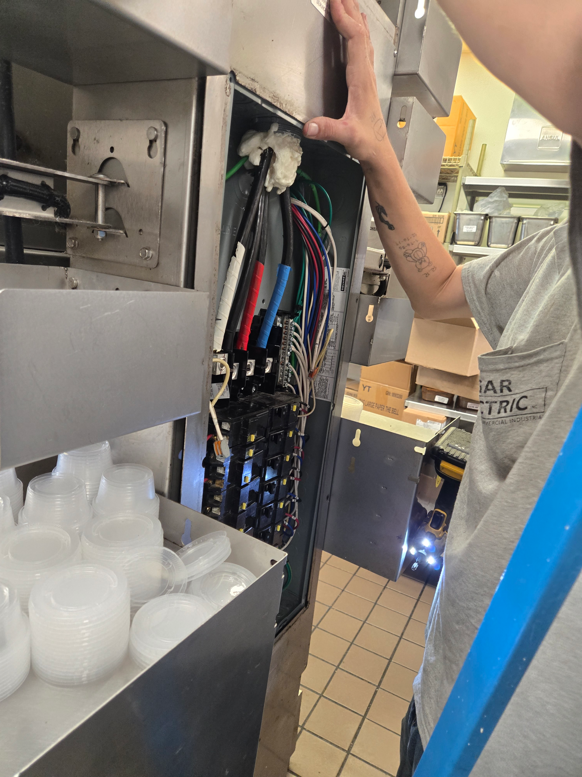 Worker inspecting an open machine panel with exposed wires in a stainless-steel kitchen area