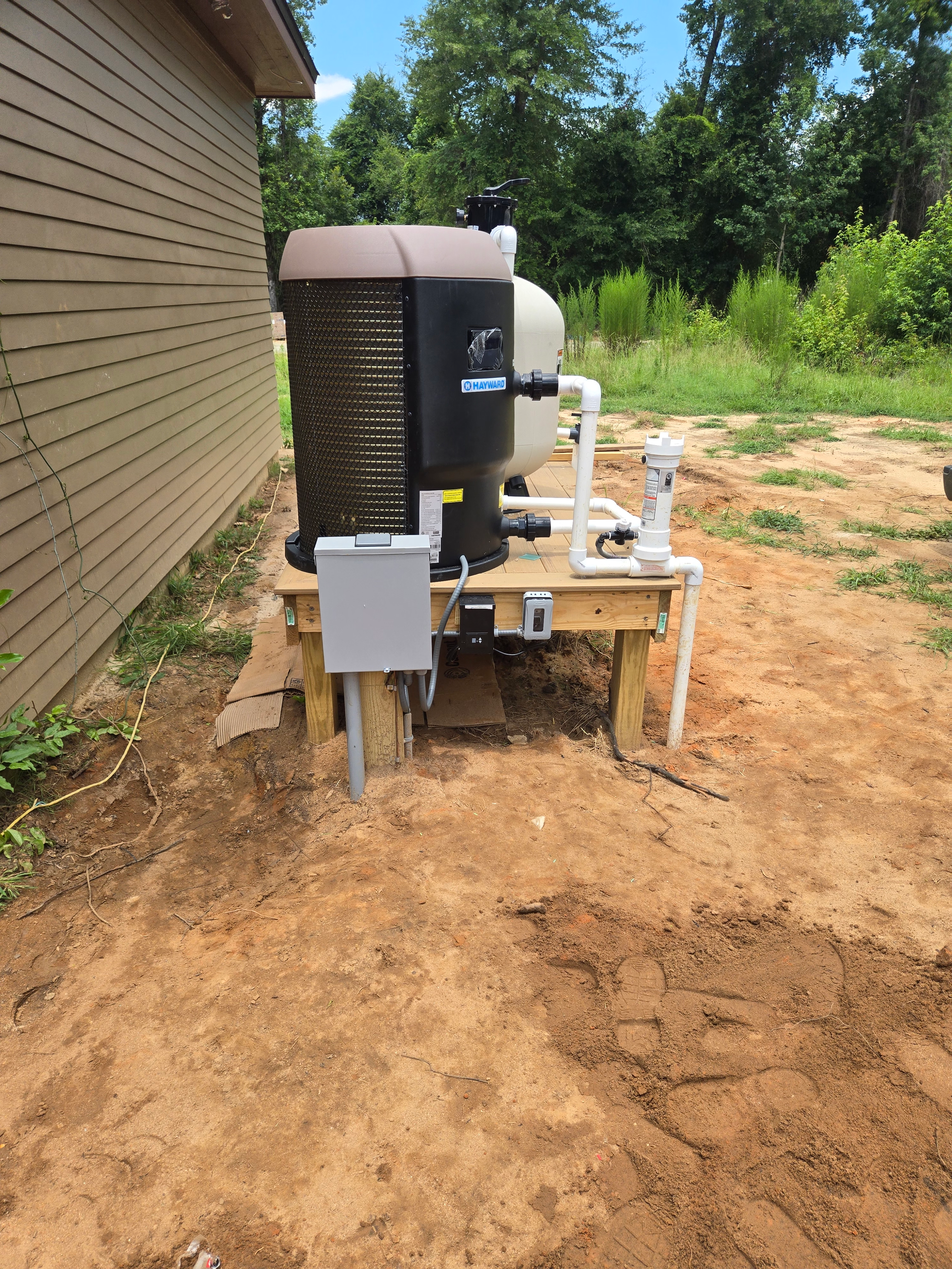 Outdoor HVAC unit beside a house, with white piping and a dirt yard under bright sunlight