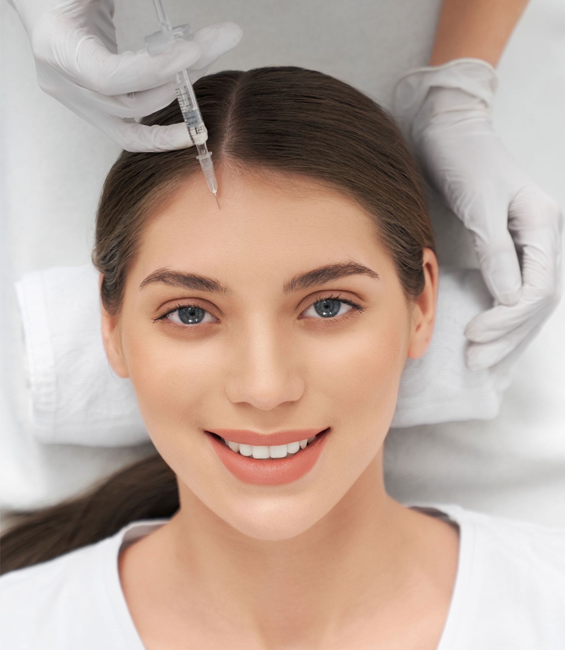 A woman is smiling while getting an injection in her forehead.